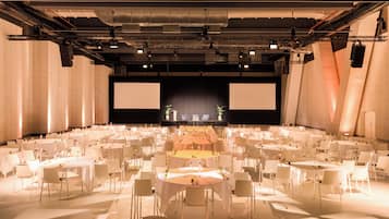 A large conference room with multiple round tables and chairs, a stage with a large screen, and a modern industrial ceiling design.