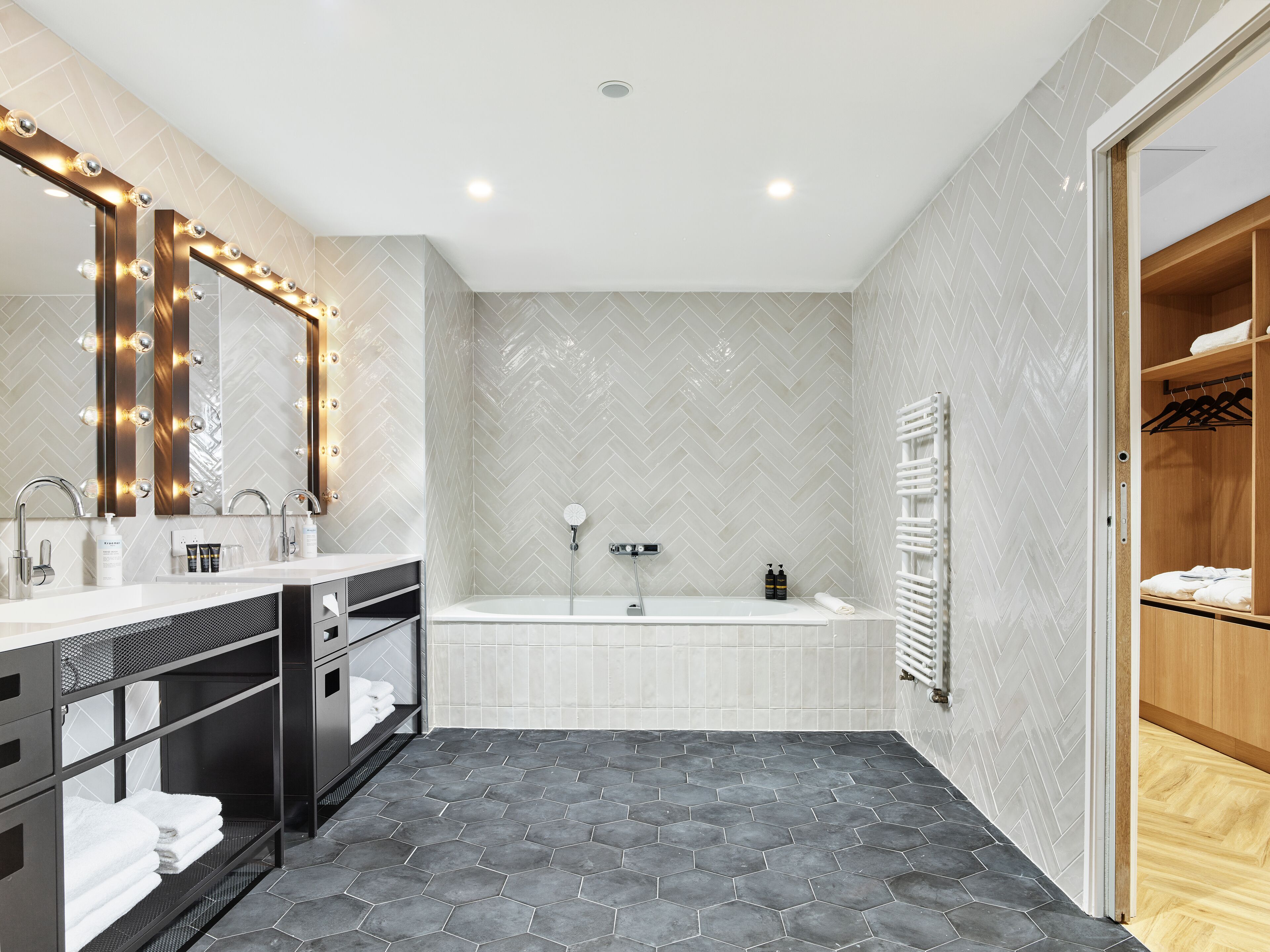 A modern bathroom with a vanity, a large mirror with lights, a bathtub, and a walk-in closet.