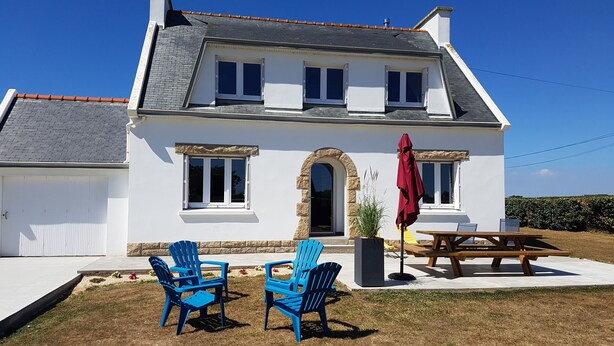 Une maison à deux étages, avec une façade blanche, une entrée surmontée d’un arc en pierre et une terrasse agrémentée de deux chaises bleues et d’un parasol rouge.