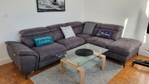 Un salon moderne avec un canapé d’angle gris, une table basse en verre, des tapis à motifs et un tableau encadré accroché au mur.