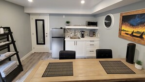 A compact kitchen with white cabinets, a refrigerator, and a microwave. There is a dining area with a wooden table and two black chairs. A painting of whales is on the wall, and a small potted plant is on the table. - Private & cozy suite located in Fort Langley (Langley Township)
