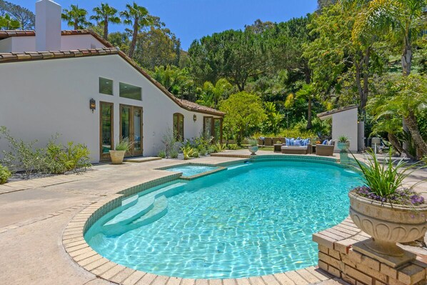 A swimming pool with a tiled edge, surrounded by a patio area with potted plants and a small structure.