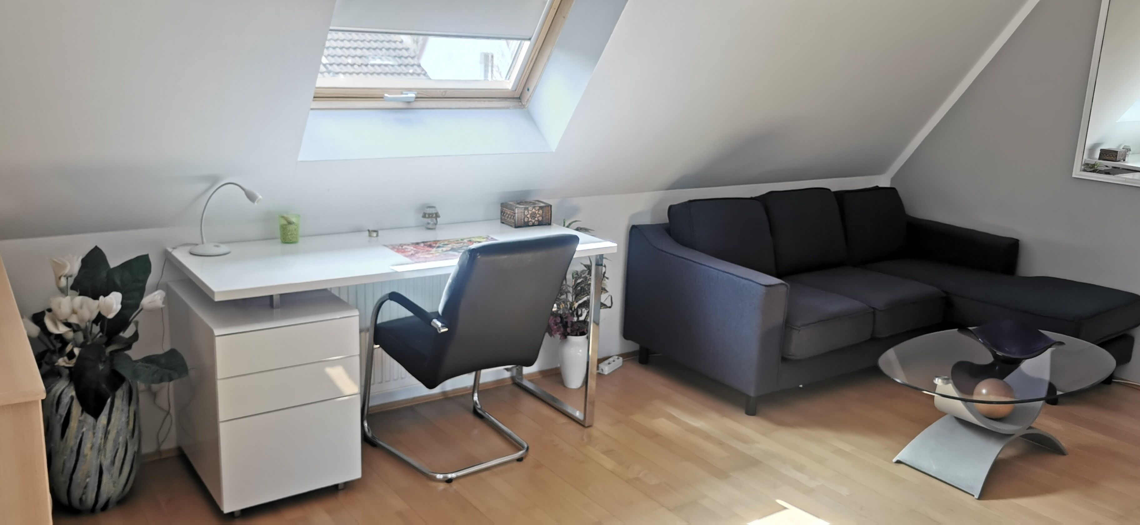 Ein modernes Schlafzimmer im Loft-Stil mit Dachfenster, Schreibtisch, einer bequemen grauen Couch und einem Glastisch.