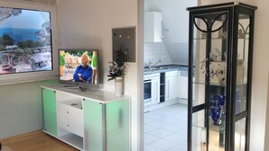 A modern living room with a TV, a balcony with a view of the sea, a kitchen area, and a glass cabinet with decorative items. - München Waldperlach Ruhig und Großzügige Wohnung (München)