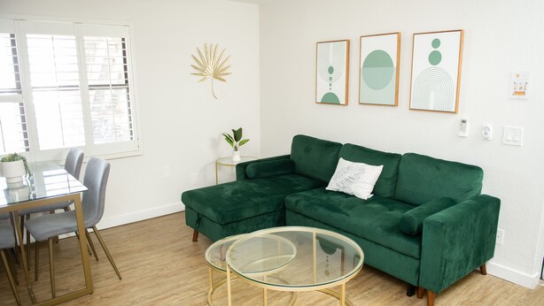 A living room with a green sectional sofa, a glass coffee table, a dining table with chairs, and a window with blinds. - Sunburst Inn The Beachside Suite - Room 5 (Indian Shores)