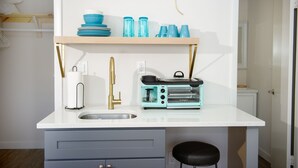 Uma cozinha compacta com bancada branca, pia, máquina de café, torradeira e prateleira com louças e copos azuis.
