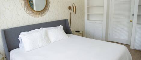 A neatly made bed with white linens, a round mirror on the wall, and a bedside table with a lamp.