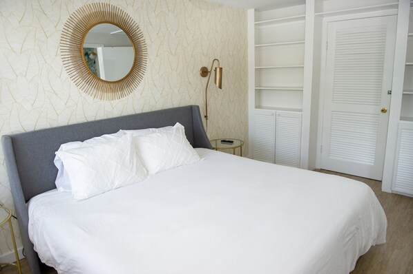 Een net opgemaakt bed met witte lakens, een ronde spiegel aan de muur en een nachtkastje met een lamp.