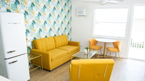 Uma sala de estar moderna com um sofá amarelo, uma mesa de centro e um espaço de jantar com cadeiras laranja. Há uma geladeira branca e um papel de parede estampado.