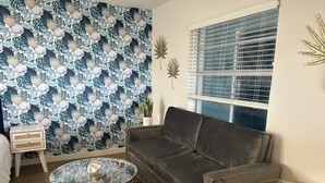 A living room with a grey sofa, two glass coffee tables with gold frames, a patterned wallpaper, a window with blinds, and a small white nightstand with a drawer. - Sunburst Inn The Beachside Classic - Room 4 (Indian Shores)