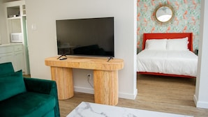 Een kamer met een televisie op een houten standaard, een groene bank, een bed met een rode hoofdeinde, en een muur met een ronde spiegel.