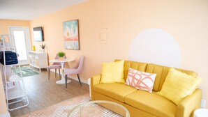 Ein Wohnzimmer mit einem gelben Sofa, einem runden Glastisch, einem weißen Sessel, einem kleinen Tisch mit Pflanze und einer weißen Wand mit großem weißen Kreis.
