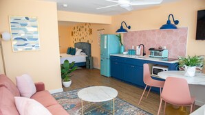 A compact living space with a kitchenette, a dining area with pink chairs, a bed with a grey headboard, and a patterned rug. - Sunburst Inn The Combo - Room 11 & 8 (Indian Shores)