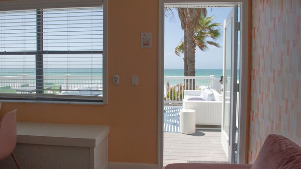 A room with a view of the ocean through a window, a door leading to an outdoor deck with a white chair and a palm tree, and a pink chair inside. - Sunburst Inn The Combo - Room 11 & 8 (Indian Shores)