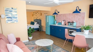 A compact living space with a kitchenette, a dining area with pink chairs, a bed with a grey headboard, and a patterned rug. - Sunburst Inn The Beachfront Suite - Room 11 (Indian Shores)