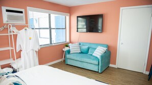 Een slaapkamer met een bed, een blauwe bank, een televisie, een raam met zonwering, een witte deur en een witte muur.