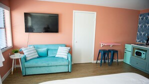 Een kamer met een blauwgroene bank, een witte deur, een kleine tafel met een plant, een roze plank en een kitchenette met een magnetron.
