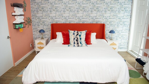 Een netjes opgemaakt bed met een rood hoofdeinde, witte beddengoed en twee rode kussens. Er staan twee nachtkastjes met lampen, een wandplank met handdoeken, en er is een patroon op de muur achter het bed.