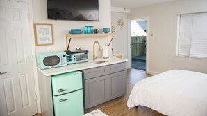 Eine kompakte Küche mit Mikrowelle, Toaster und Spüle. Es gibt einen kleinen Kühlschrank, ein weißes Bett und einen an der Wand montierten Fernseher.