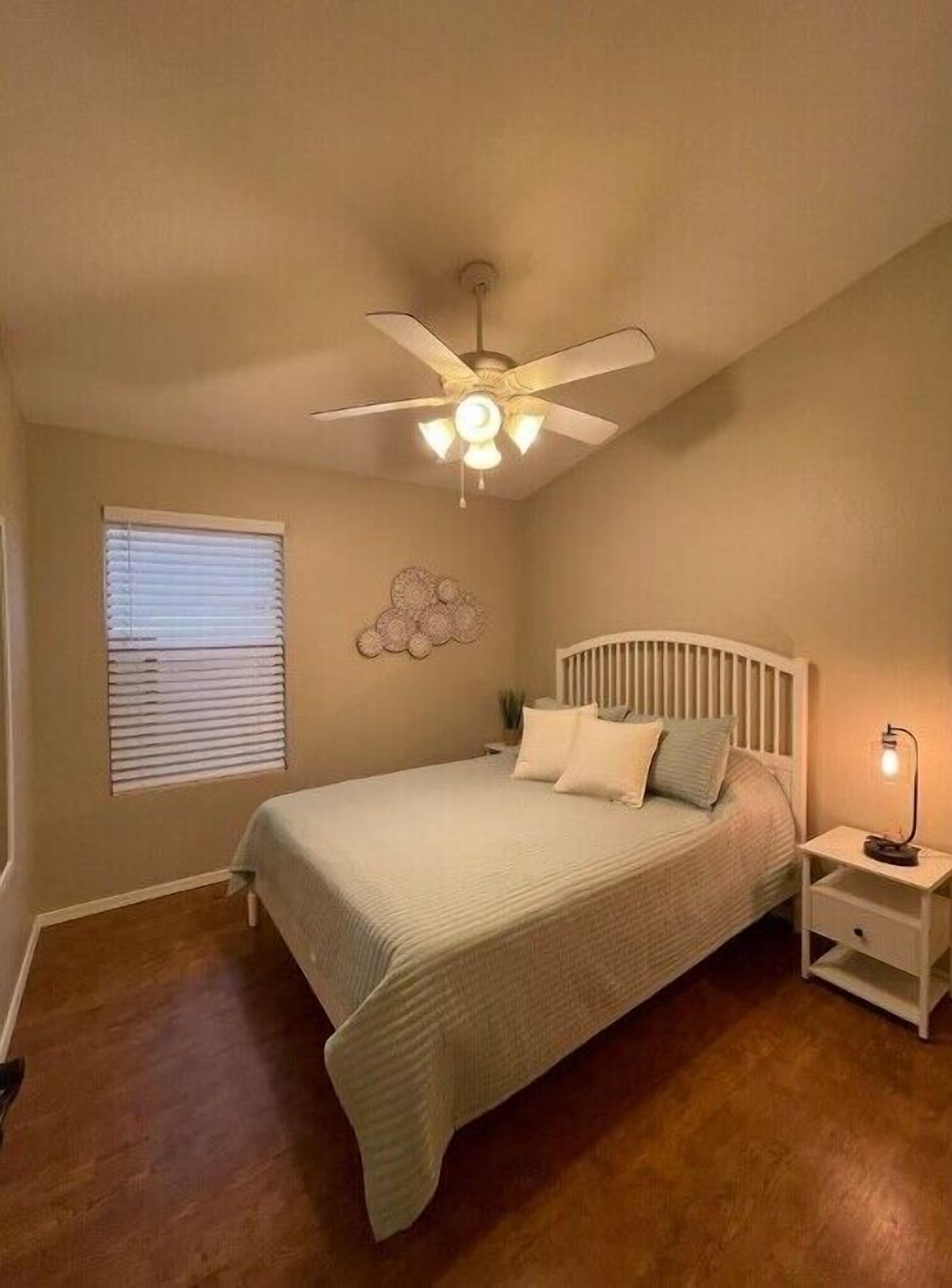 Une chambre à coucher comprenant un lit, un ventilateur de plafond, une table de chevet et un luminaire fixé au mur.
