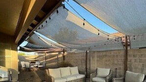 Een terras buiten met een eethoek, rieten meubels en een vloerkleed met een tropisch bladmotief.