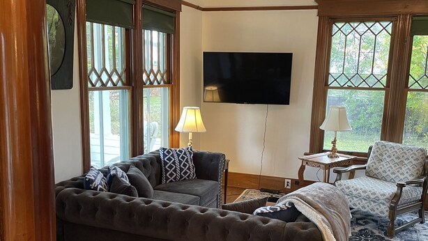 A living room with a dark-colored sofa, a television mounted on the wall, and a patterned armchair. - Historic 2 bedroom home in downtown Stevensville. Pet friendly! RV parking (Stevensville)