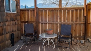 A wooden deck with two wicker chairs and a round table, a potted plant, and a window on the left. - Mt Williamson Motel and Basecamp (Independence)
