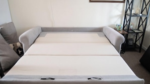 A neatly made bed with a grey headboard and footboard, a small bedside table with a decorative item, and a black shelving unit against the wall. - Mt Williamson Motel and Basecamp (Independence)
