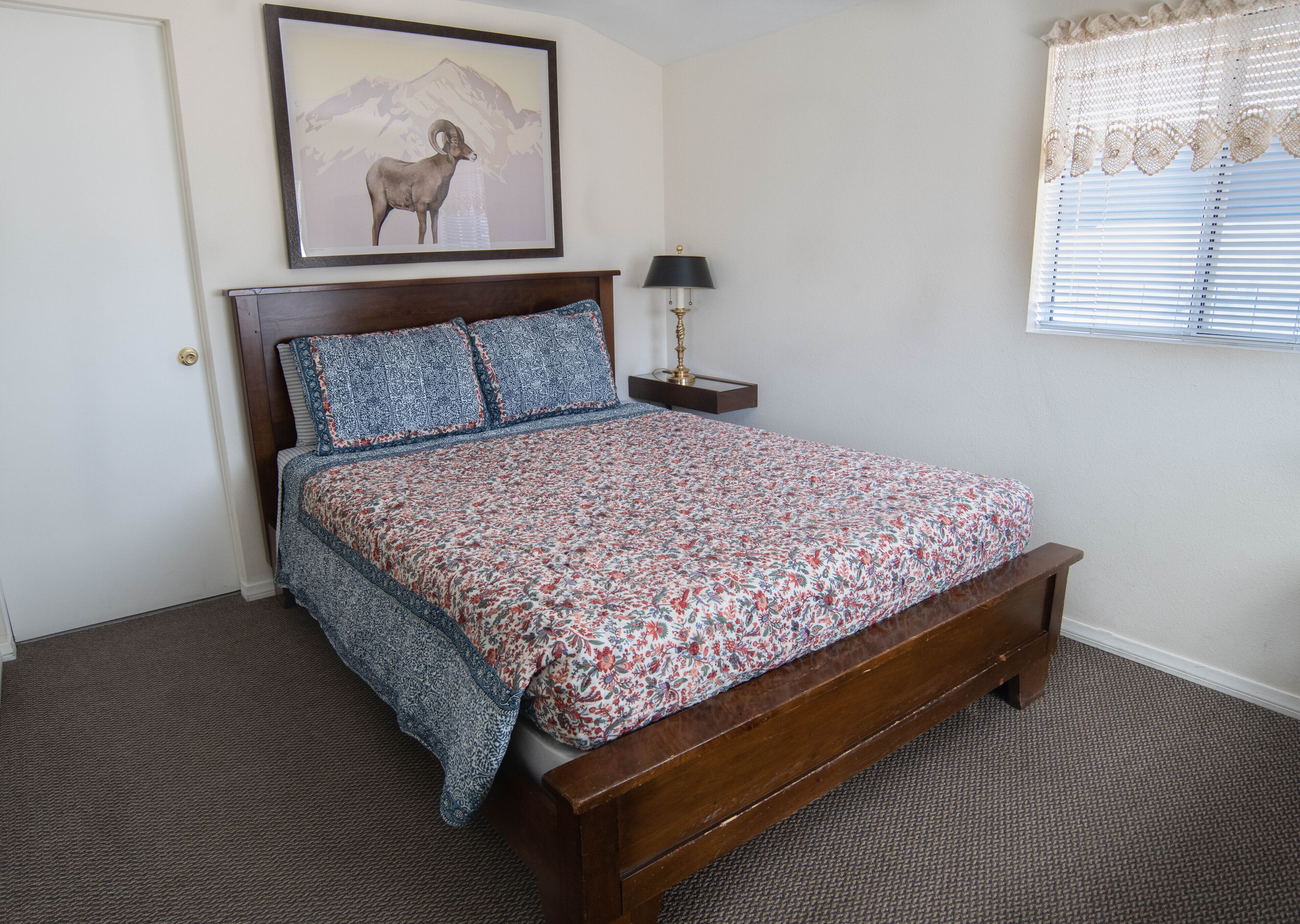 A bedroom with a wooden bed, floral bedding, a framed picture on the wall, a nightstand with a lamp, and a window with blinds.