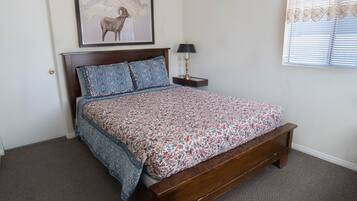 A bedroom with a wooden bed, floral bedding, a framed picture on the wall, a nightstand with a lamp, and a window with blinds.