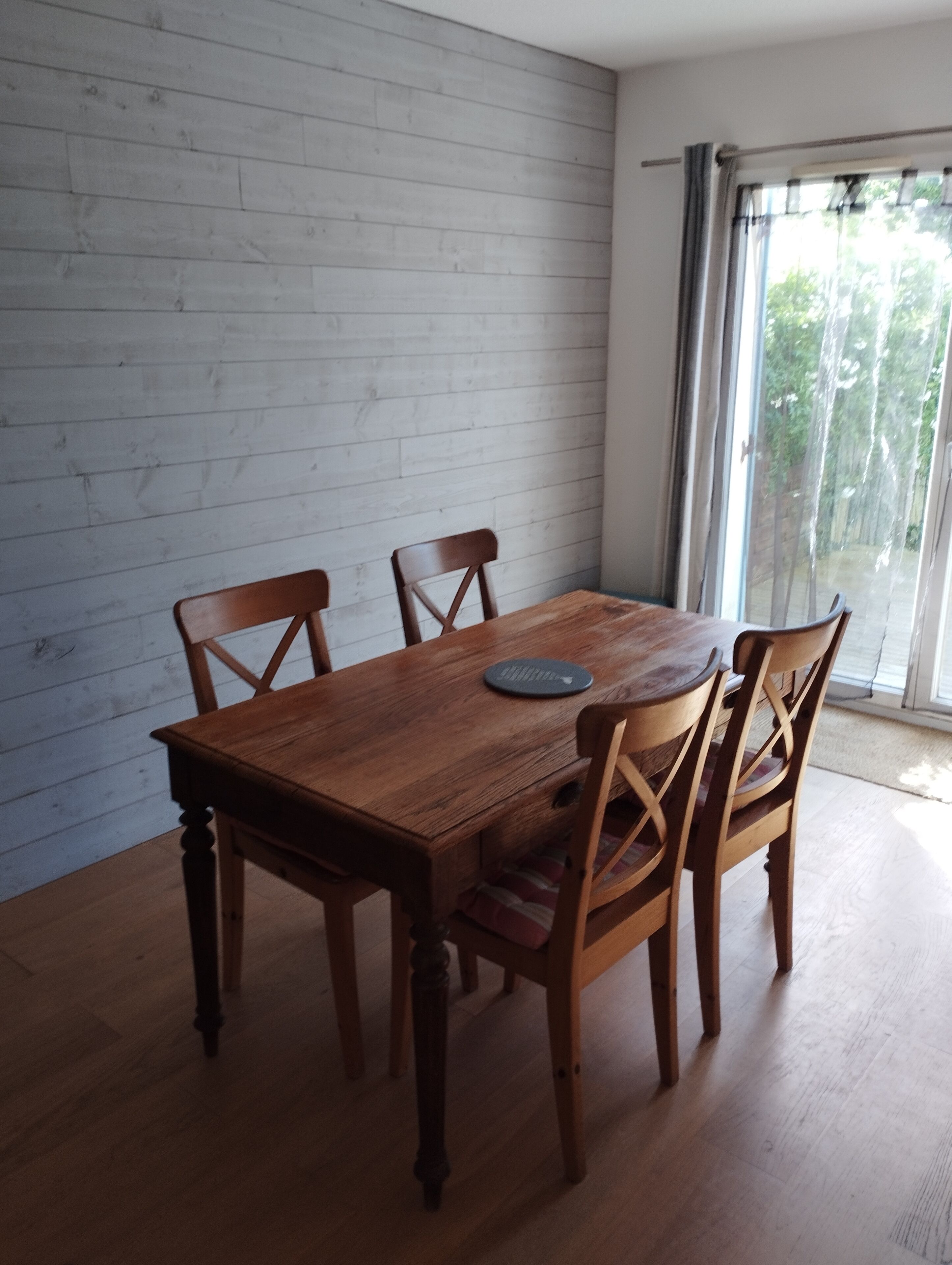 A dining area with a wooden table and chairs, a round table runner, and a view through a window with curtains.