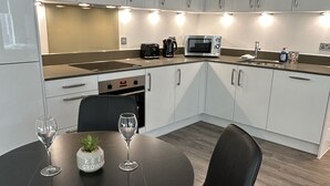 Una cocina moderna con armarios blancos, un microondas, una cafetera y un fregadero. Hay una mesa de comedor con dos sillas y una pequeña planta en maceta.