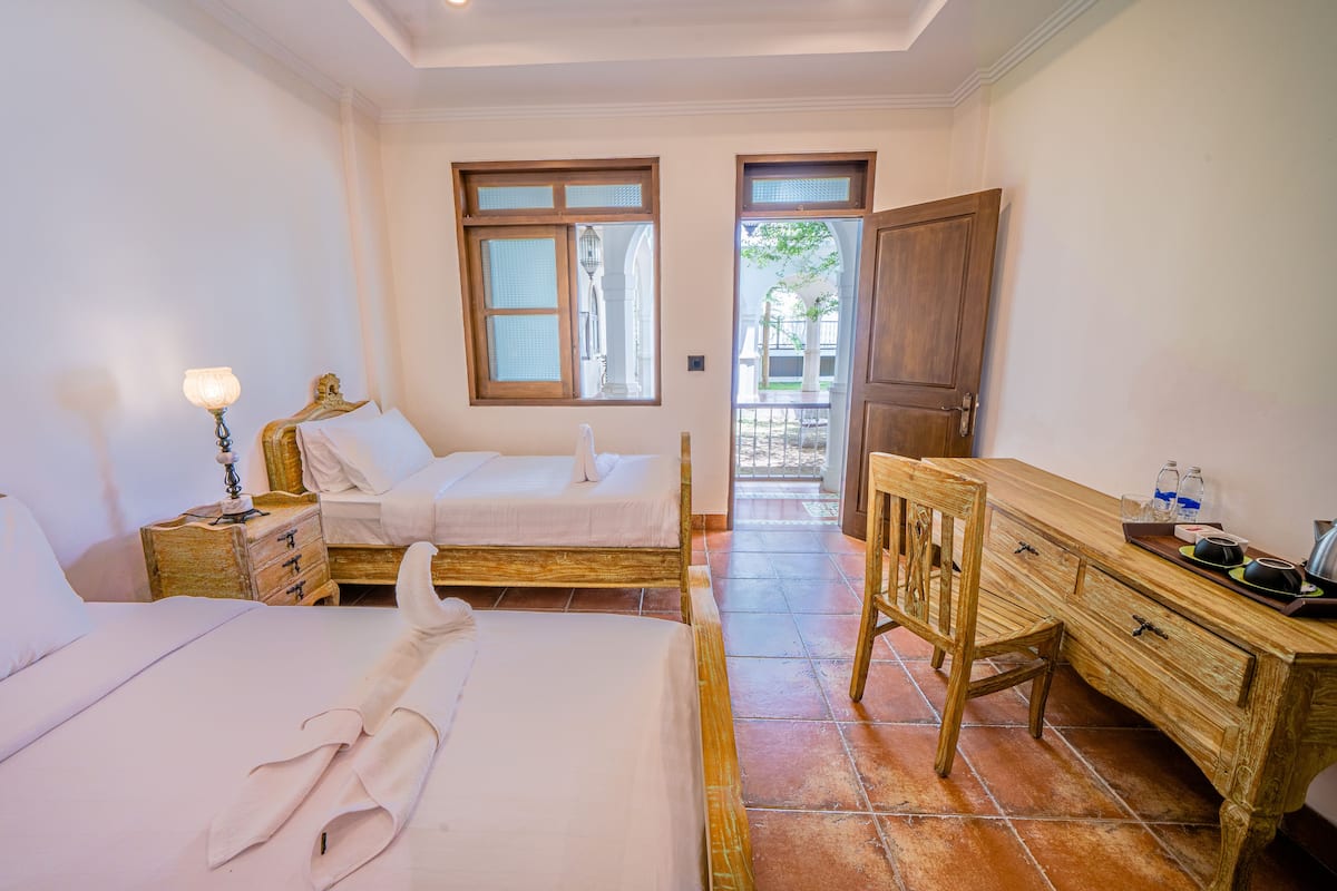 Une chambre avec deux lits, une commode en bois, une table de chevet avec une lampe, et une vue sur l’extérieur par une porte.