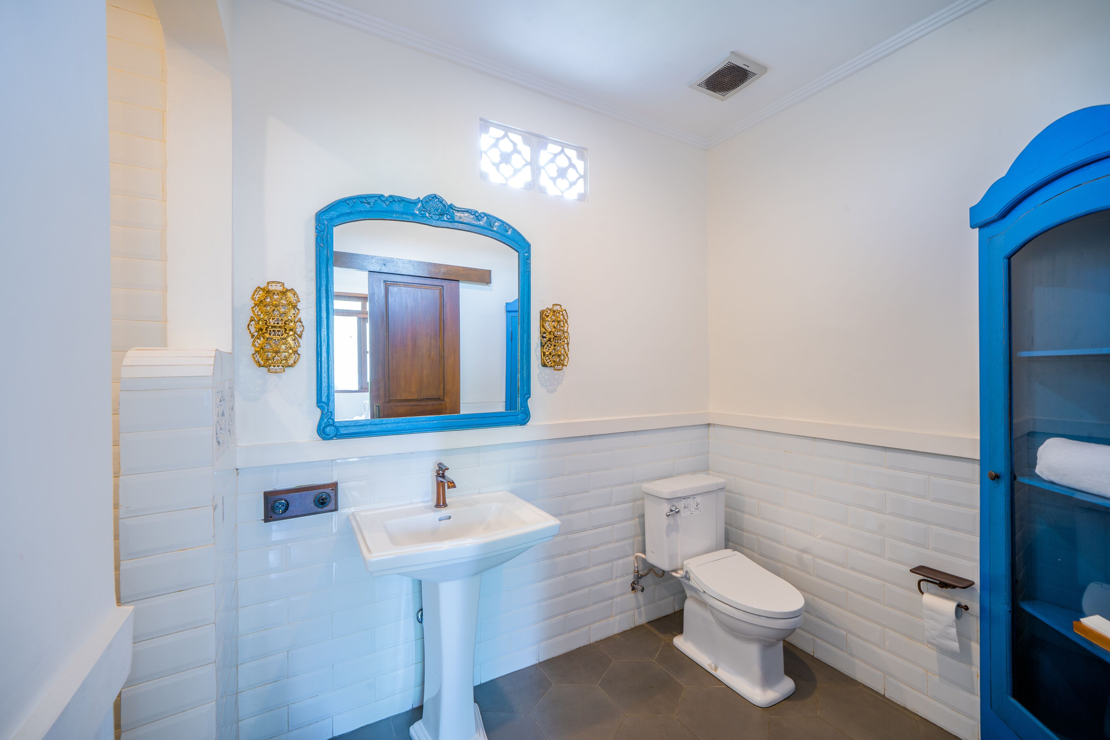 A bathroom with white tiled walls, a white sink, a blue-framed mirror, a toilet, and a blue cabinet with shelves.