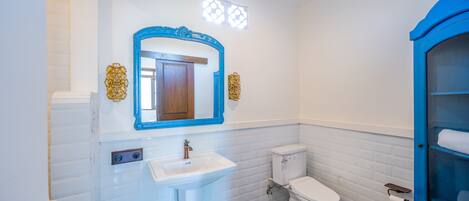 A bathroom with white tiled walls, a white sink, a blue-framed mirror, a toilet, and a blue cabinet with shelves.