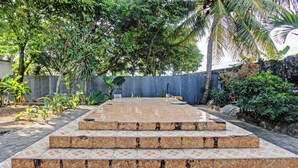 A tiled outdoor area with steps leading to a seating area surrounded by lush greenery and a corrugated metal fence. - Hotel O Losmen Tjabe Merah Near Alfamart Bintaro (Purwokerto)