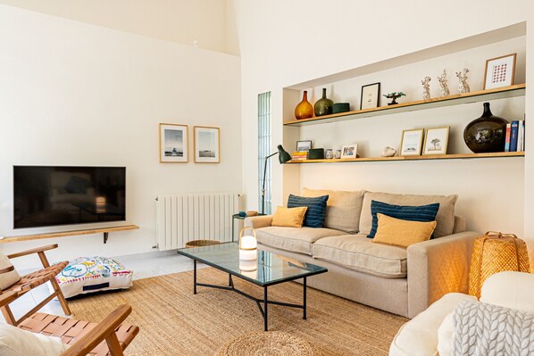 A living room with a sofa, coffee table, and TV. There are shelves with decorative items and a lamp.