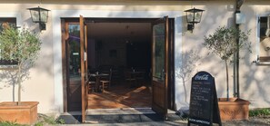 L’entrée du restaurant est ouverte, et on peut voir à l’intérieur un sol en bois, des tables et des chaises. À l’extérieur, il y a des plantes en pot, et un tableau noir affiche le menu sur le côté droit.
