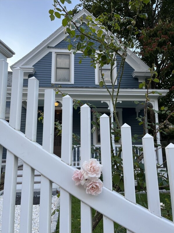 Una cerca blanca de piquetes con flores rosadas frente a una casa azul con porche.