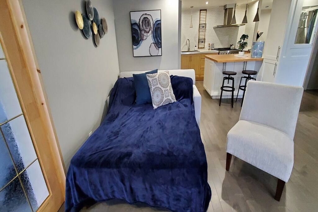 A bedroom with a bed, a white chair, and a kitchenette in the background.