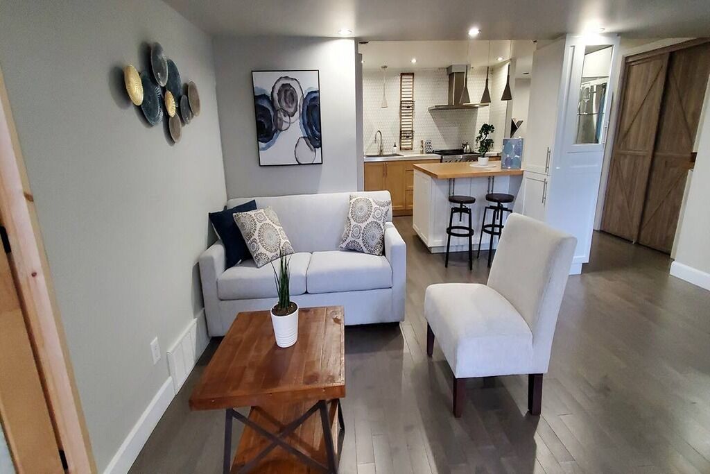 A modern living room with a sofa, armchair, and coffee table. A kitchen is visible in the background with bar stools.