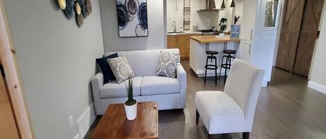 A modern living room with a sofa, armchair, and coffee table. A kitchen is visible in the background with bar stools.
