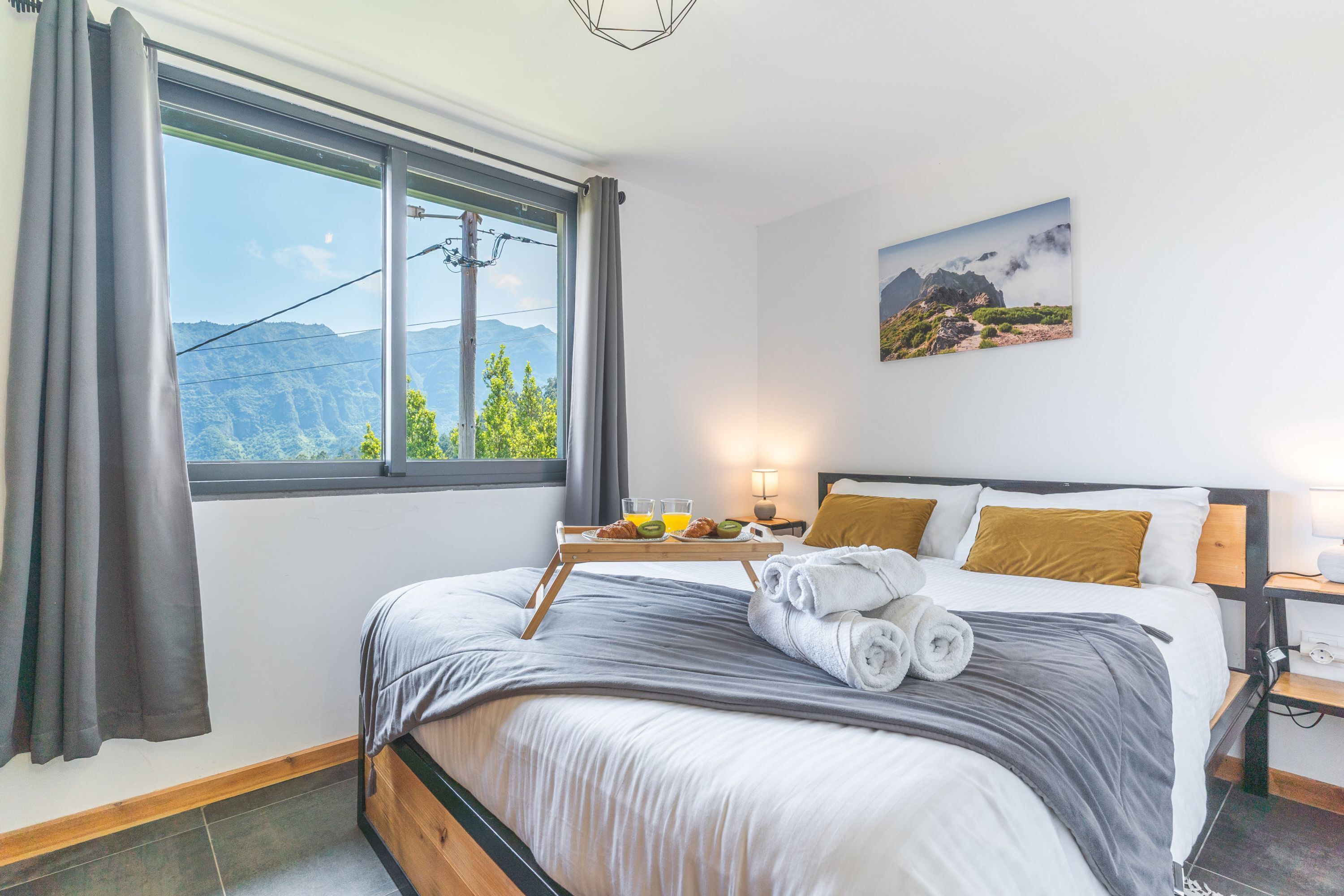 A neatly made bed with a grey and white duvet, a tray with breakfast items, and a mountain view through the window.