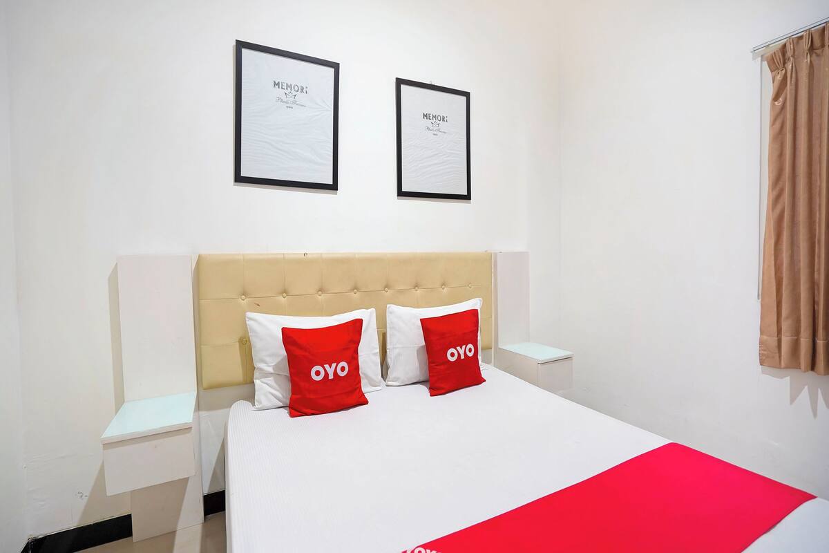 A hotel room with a bed, two framed pictures on the wall, a nightstand, and a window with curtains.