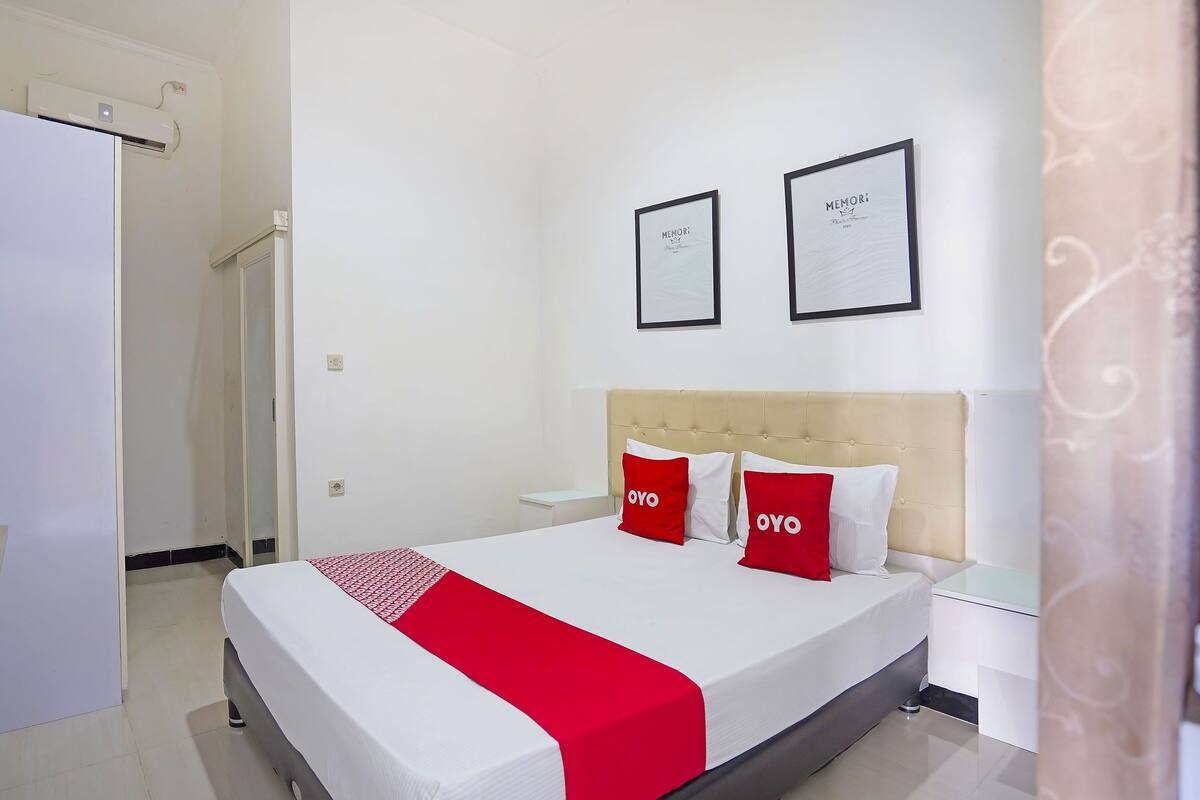 A hotel room with a bed, a nightstand, a wall-mounted air conditioner, and two framed certificates on the wall.