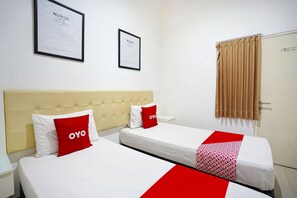Et hotelværelse med to senge, begge med hvide sengetæpper og røde puder, en beige sengegavl og to billeder i rammer på væggen.