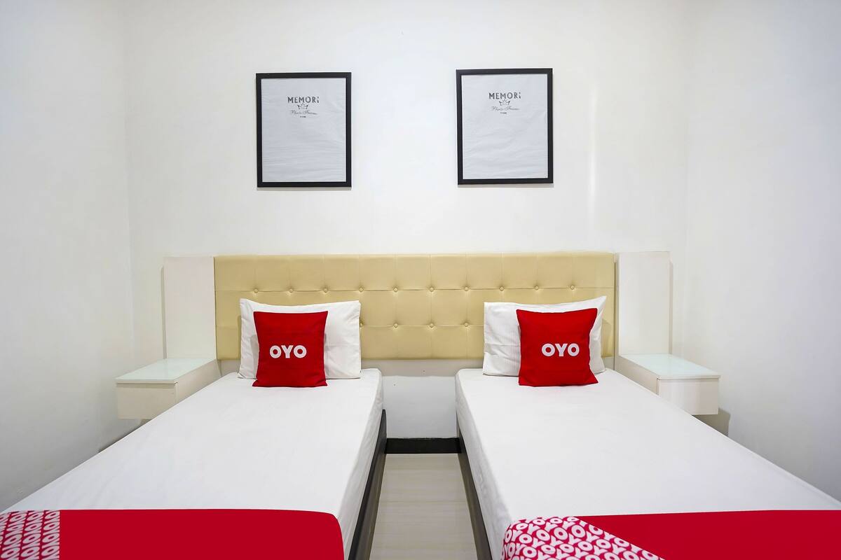 A hotel room with two beds, a tufted headboard, white bedding, red pillows, and framed pictures on the wall.