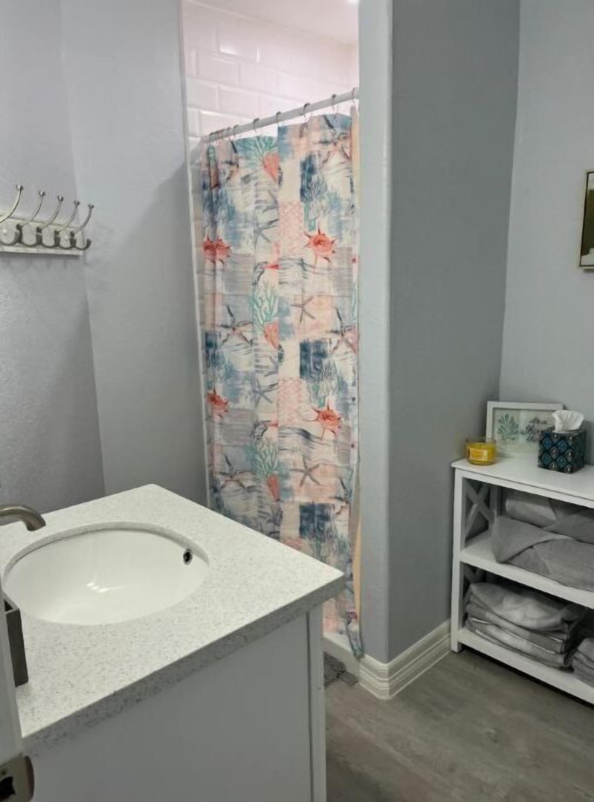 Une salle de bain avec un lavabo blanc, un rideau de douche à motifs tropicaux, une petite étagère avec des articles de toilette et un meuble blanc.