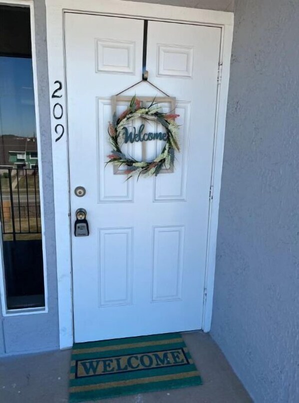 Puerta blanca con un adorno de bienvenida y un felpudo que dice 'Welcome'.