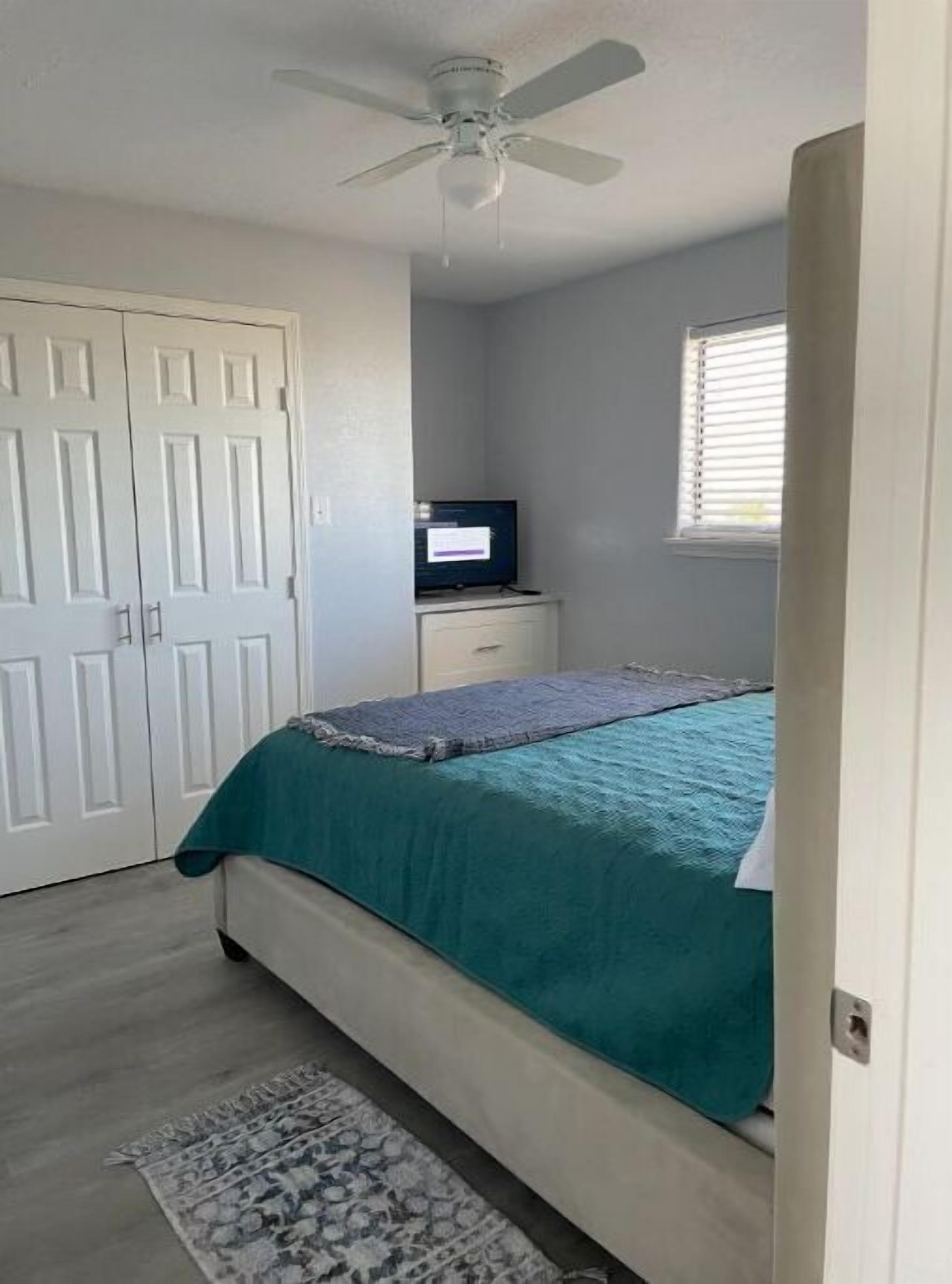 Un dormitorio con cama, ventilador de techo, televisor sobre un mueble y ventana con persianas.